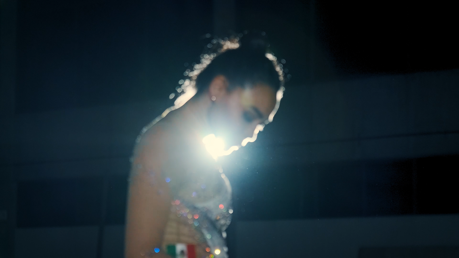 A female performer in a sparkling costume stands under a dramatic spotlight on a dark stage.
