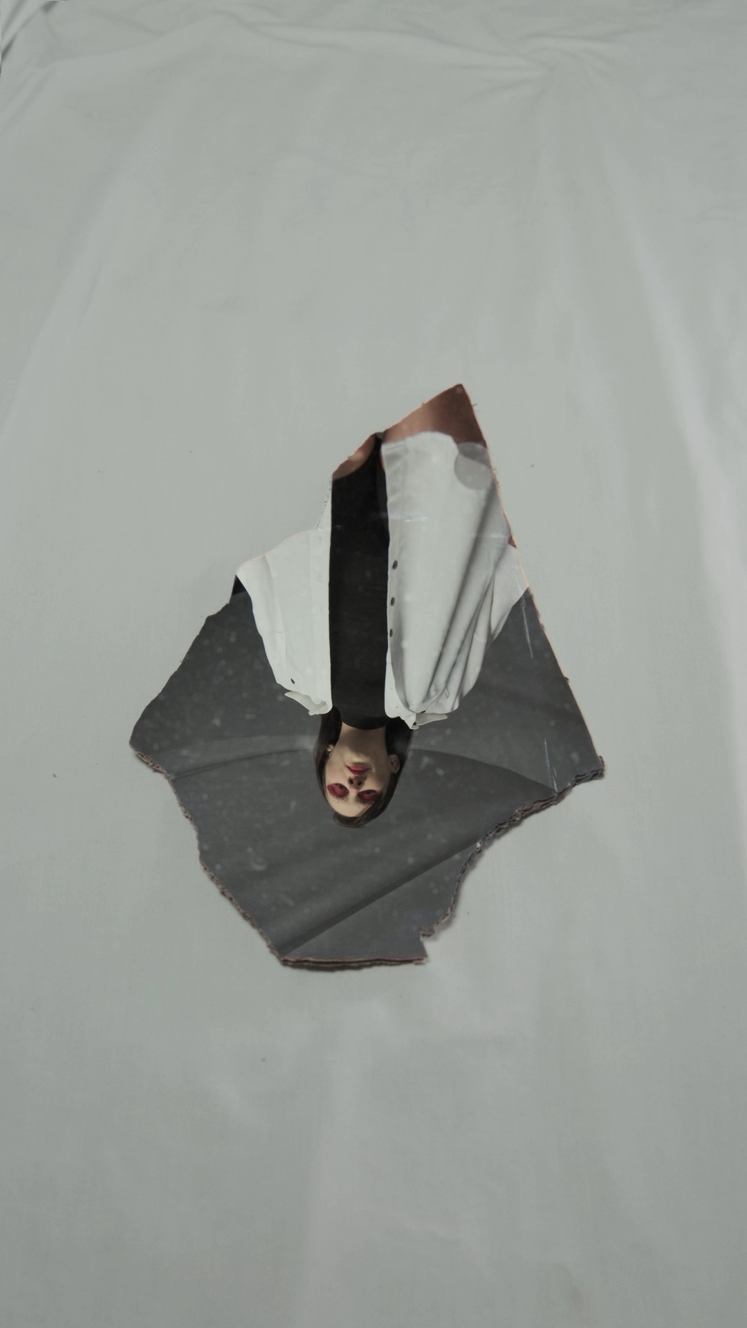 Person in white shirt and black tie emerging upside down through a torn hole in white surface.
