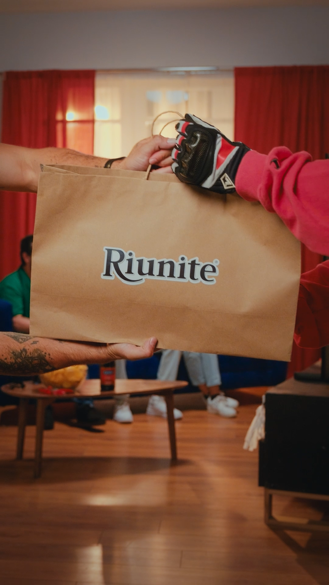 A person hands over a large brown paper bag branded with the Riunite logo in a living room.