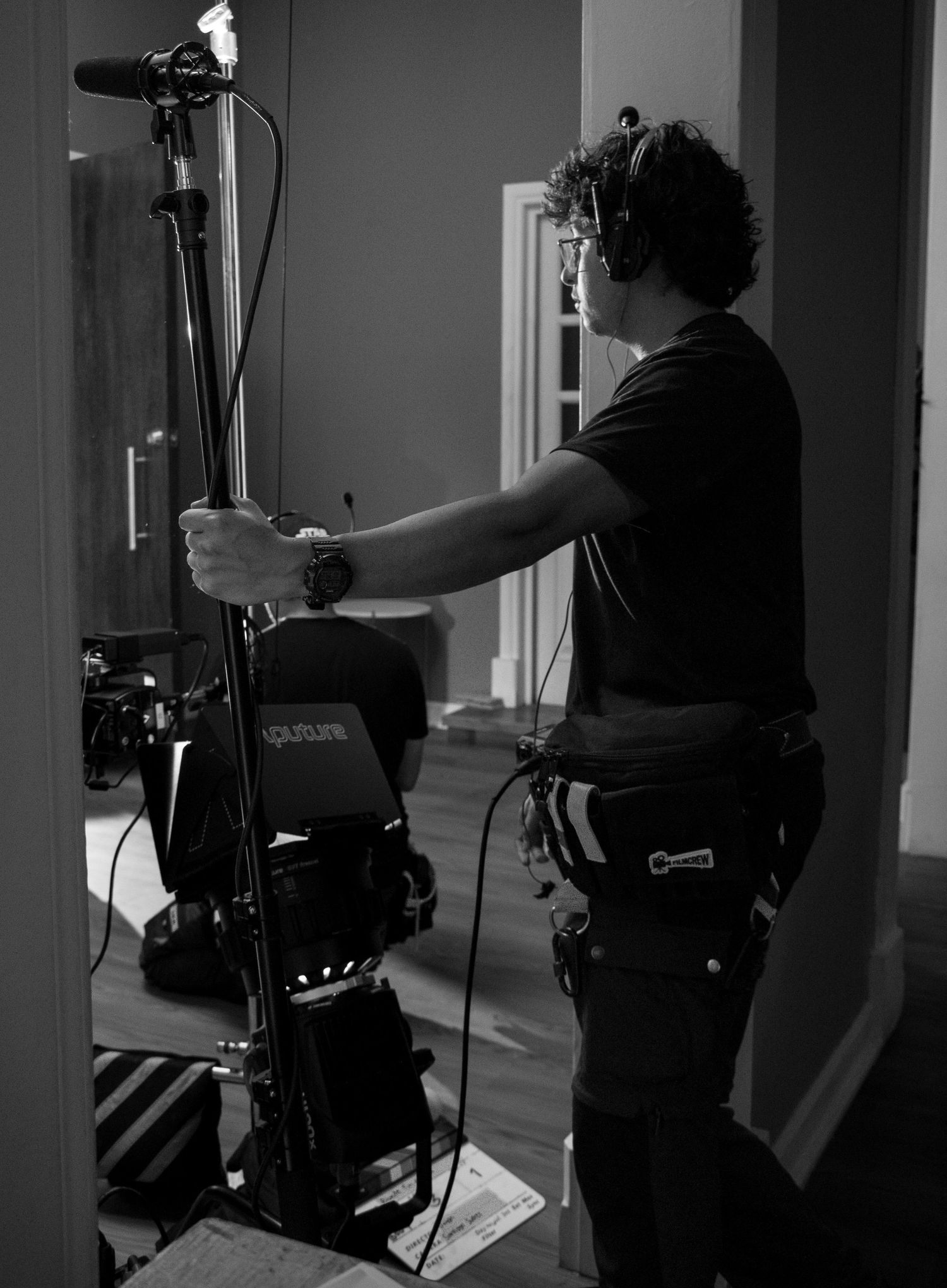 Black and white photo of boom operator holding microphone pole beside Aputure lighting equipment on set.