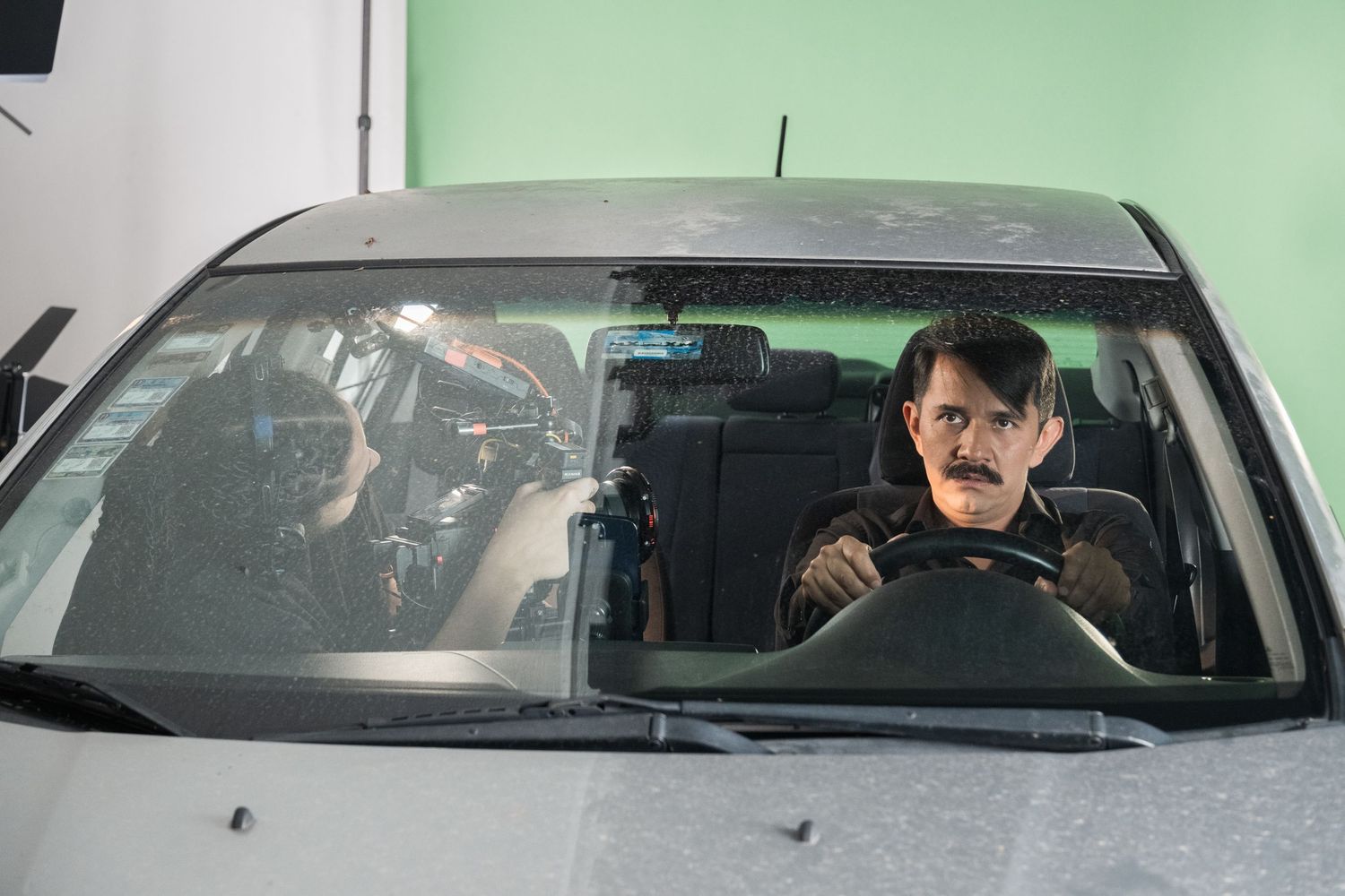 A man with a mustache sits in the driver seat of a car on a green screen film set.