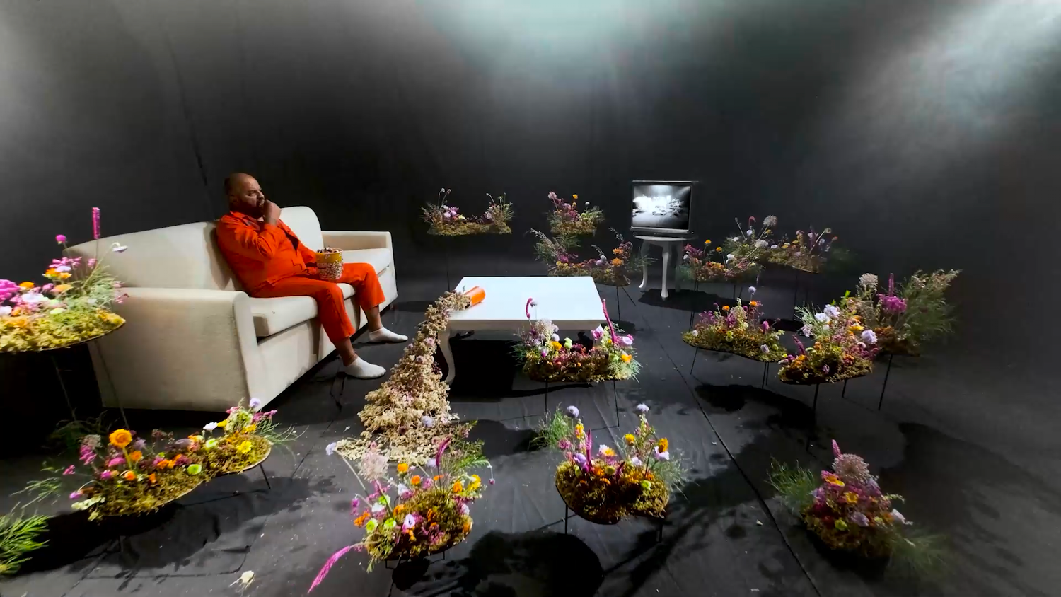 Man in orange outfit sits on white sofa surrounded by colorful flower arrangements in a dark studio setting.