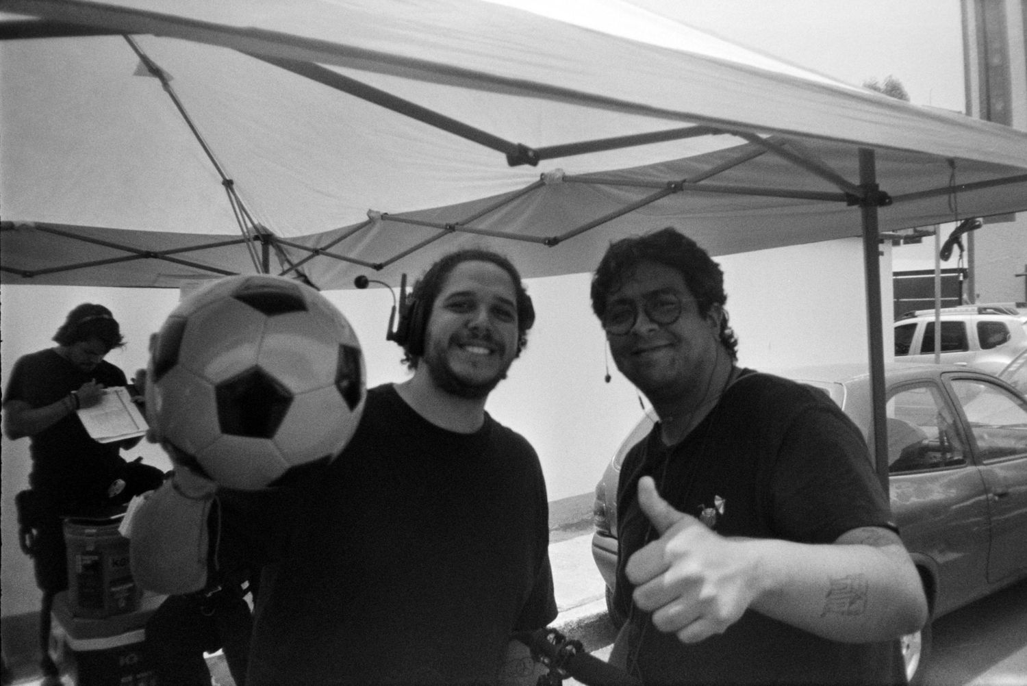 Two men smile under a tent on a film set, one giving thumbs up beside a soccer ball.