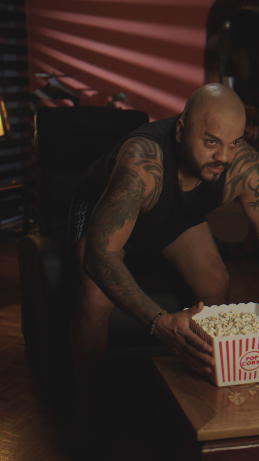 Tattooed bald man leaning over a coffee table with a bucket of popcorn in a dark room.
