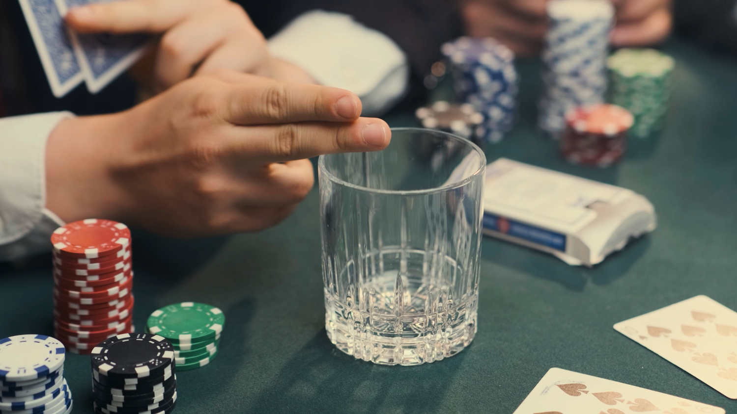 Hands holding playing cards near poker chips and a crystal whiskey glass on a green casino table.