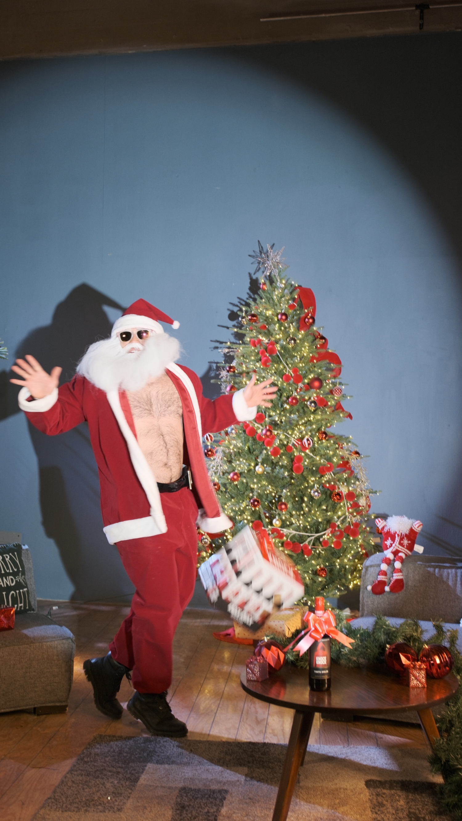 Person in Santa suit with exposed chest and sunglasses posing beside a decorated Christmas tree indoors.