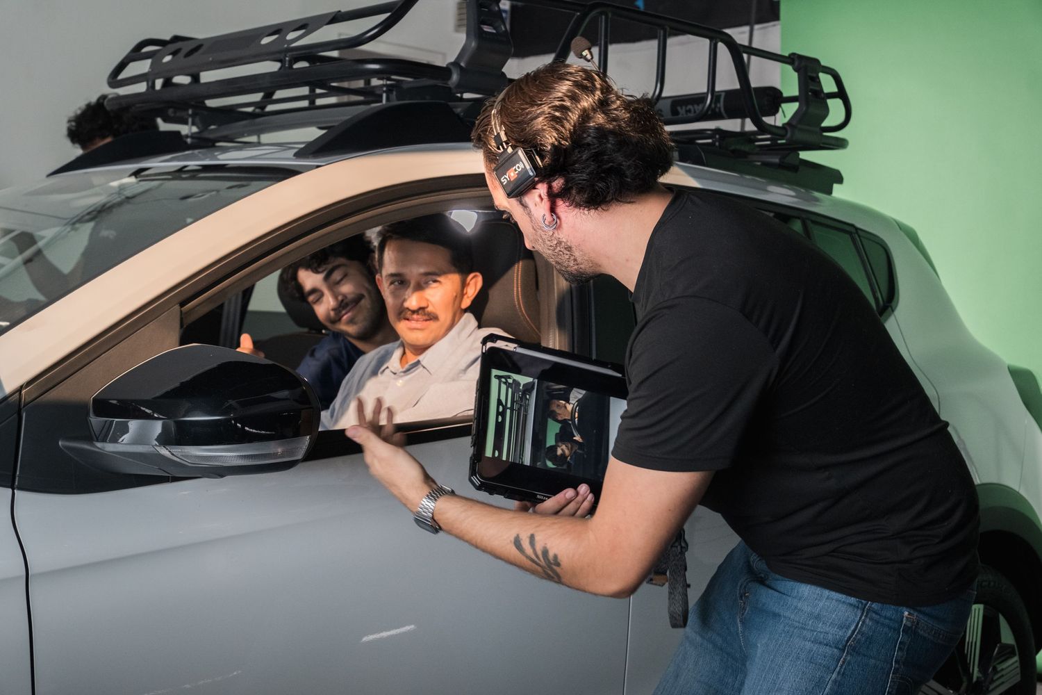 Film crew reviews footage on tablet with actors seated in car on green screen set.