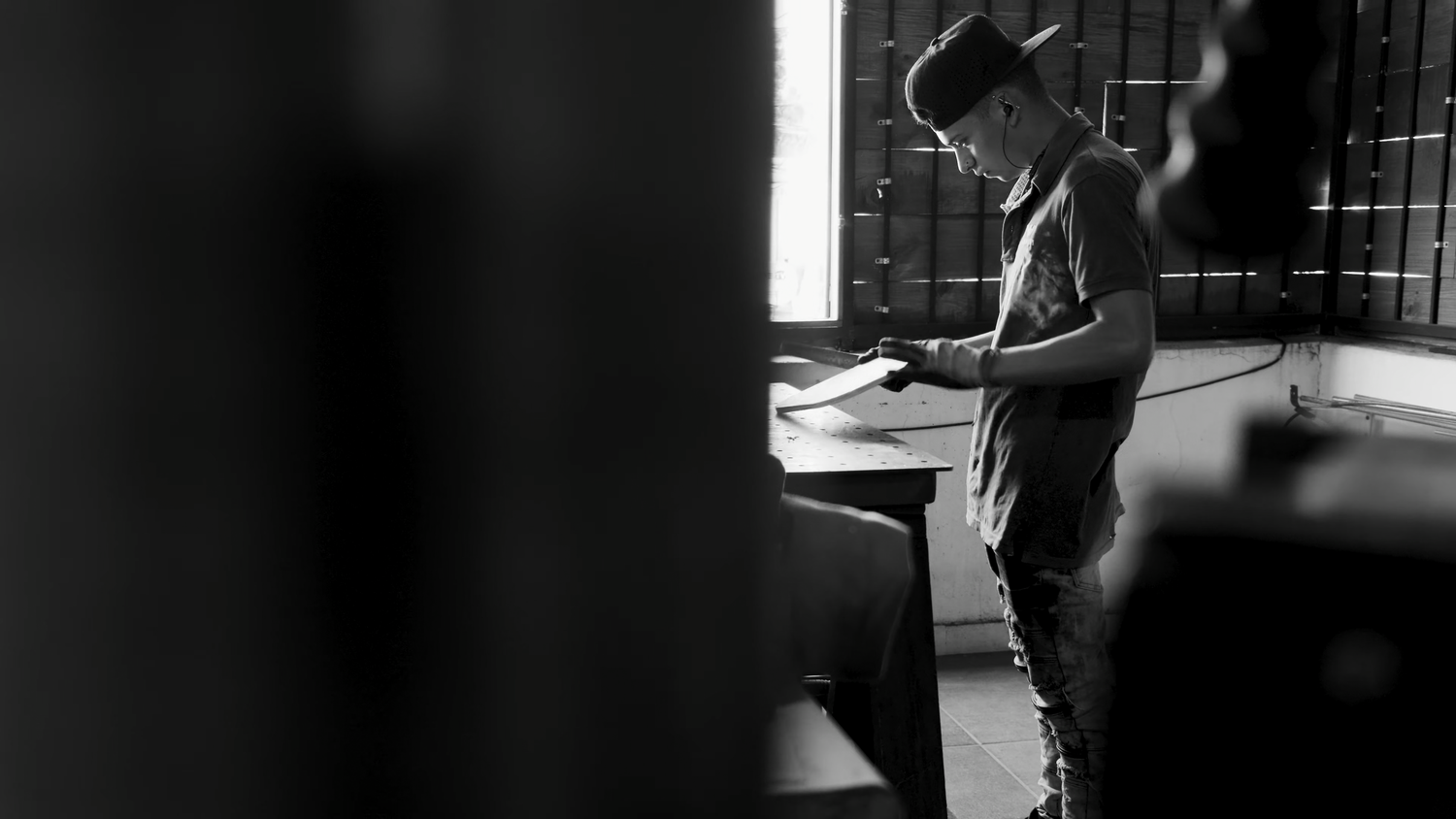 Black and white photo of a young man standing at a desk reading documents in a dimly lit room.