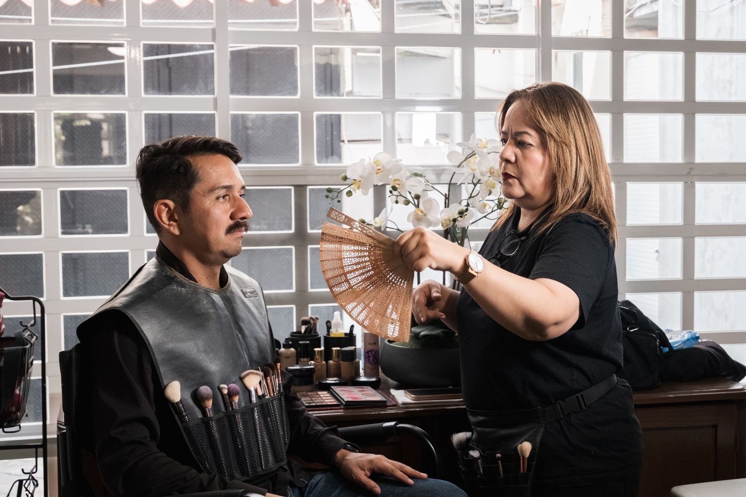 Makeup artist using a fan while applying makeup to a male actor in a salon chair.