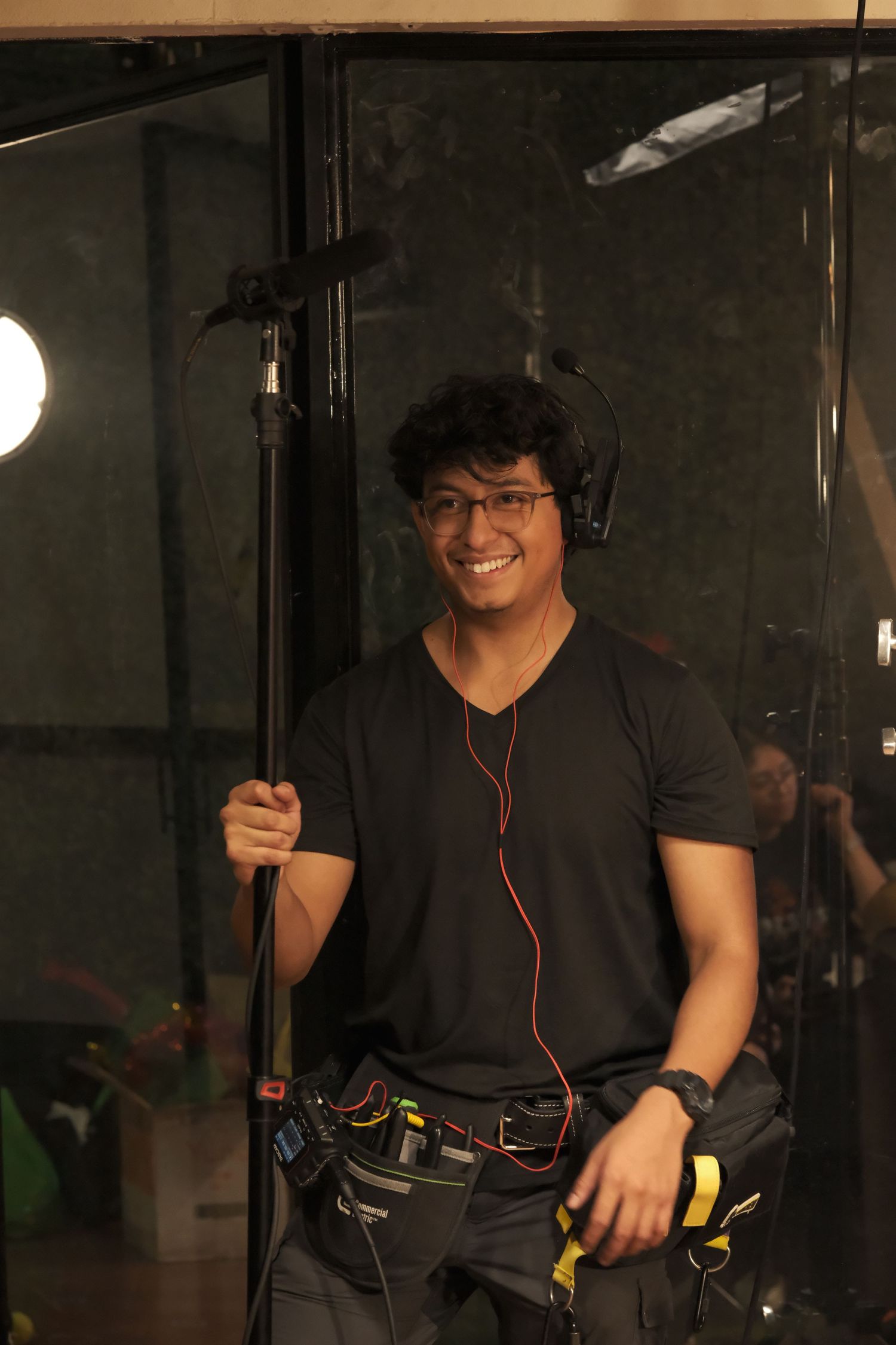 Smiling male film crew member holding a boom pole with red earphones on a professional movie set.