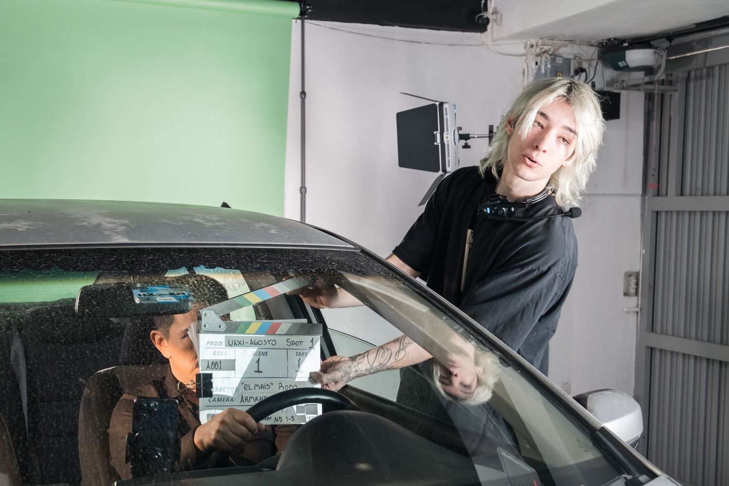 Female director with blonde hair holding a clapperboard next to a car on a green screen set.