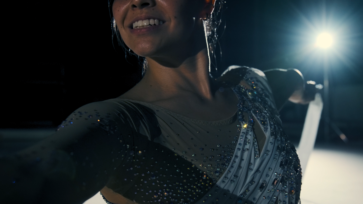Smiling female rhythmic gymnast in sparkly dark leotard performs under bright stage lighting.