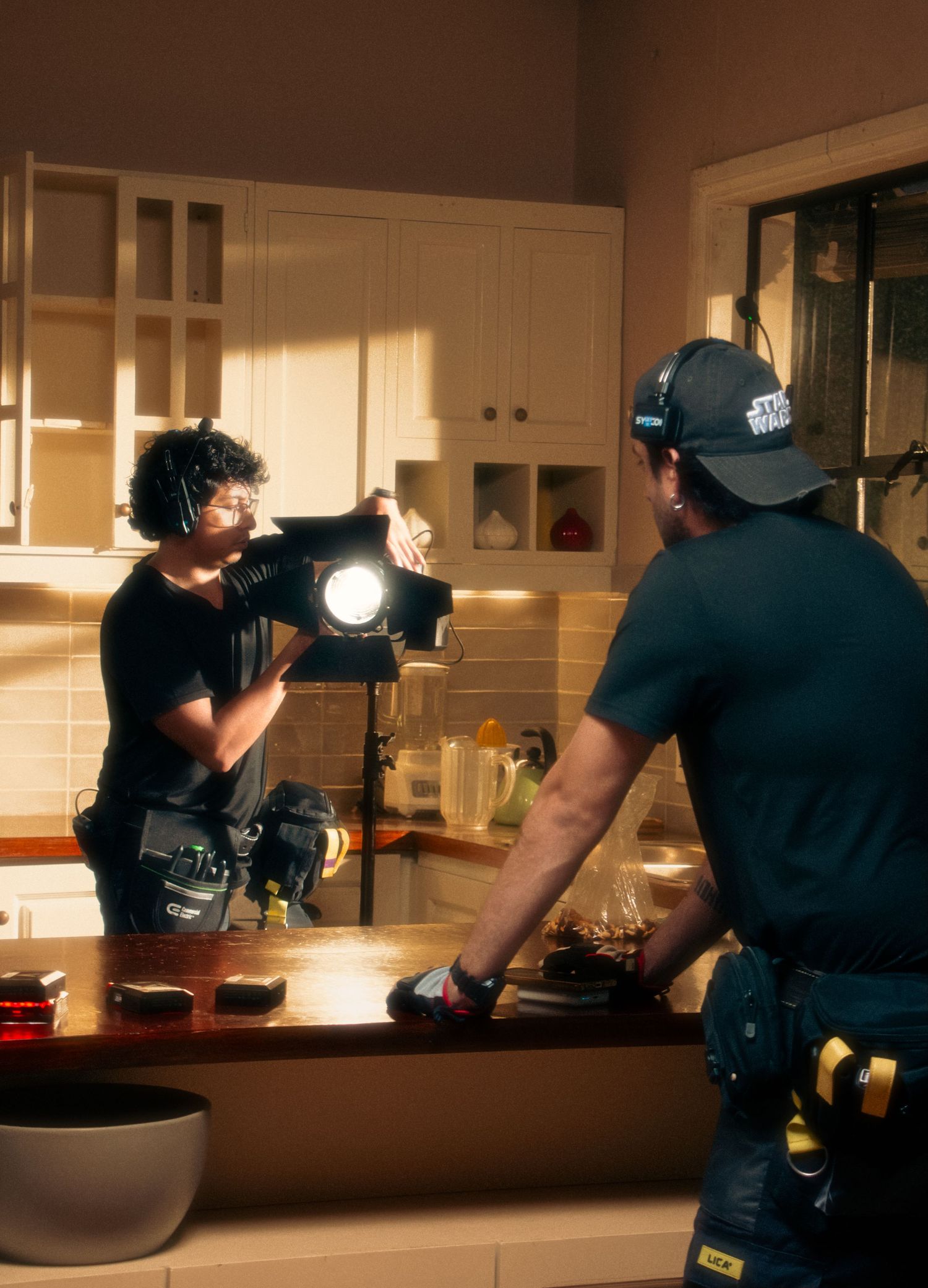 Two film crew members adjust lighting equipment in a warmly lit kitchen during production.