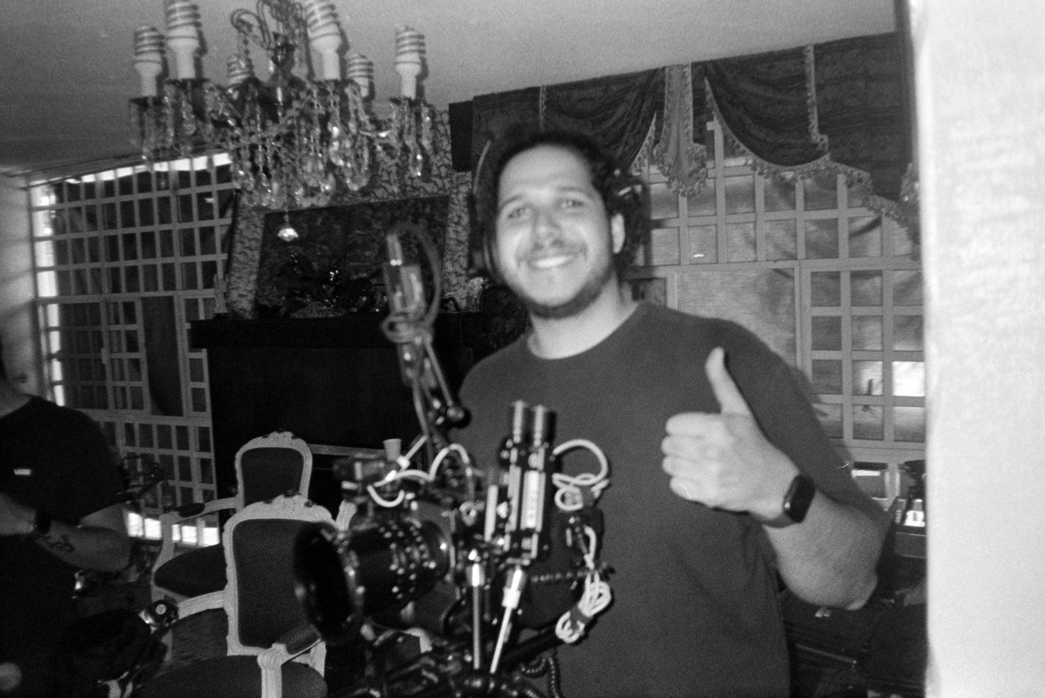 Black and white photo of smiling man giving thumbs up while holding camera rig indoors.