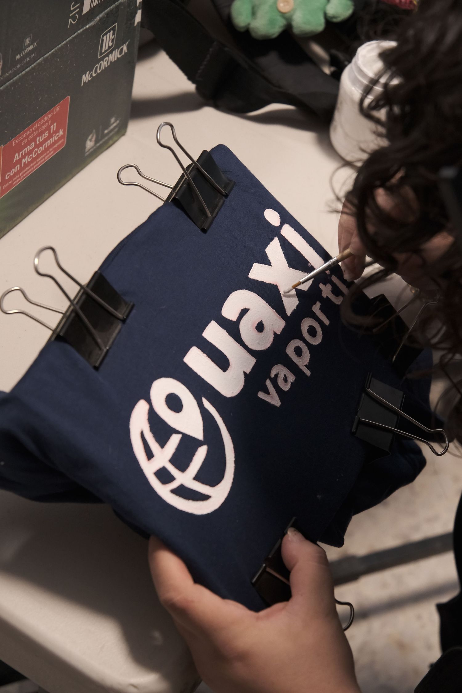 Hands holding a navy blue Quaxi Vaporti branded tote bag secured with binder clips on a white surface.