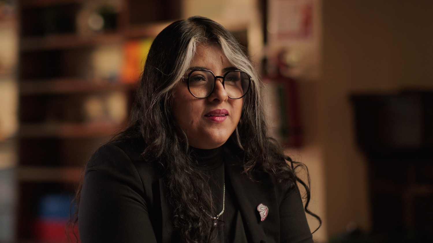 Woman with long dark hair and glasses wearing a black turtleneck speaks in a warmly lit indoor setting.