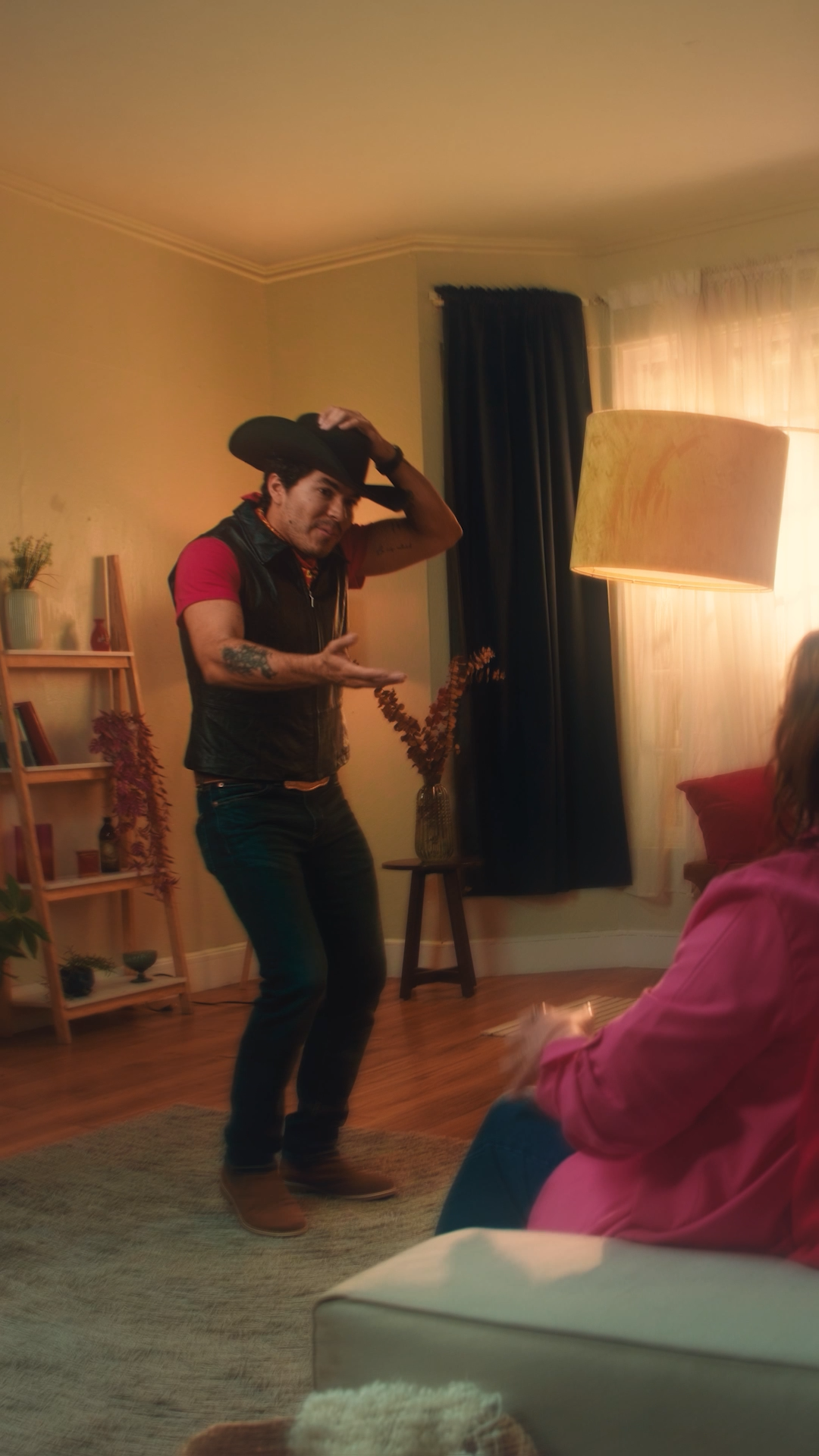 Man wearing cowboy hat and vest dancing in a living room for a woman on a couch.