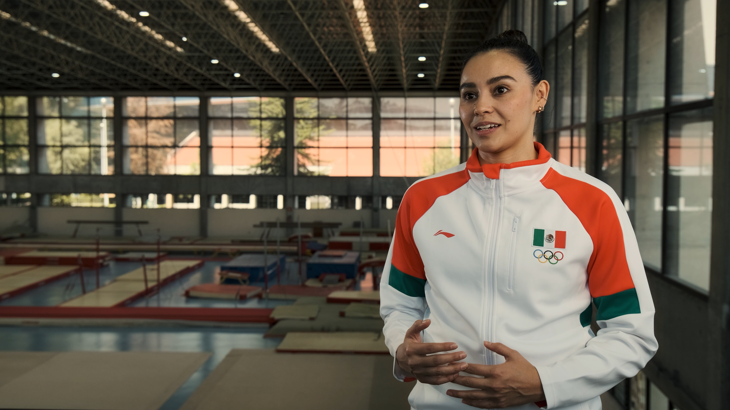 A female athlete in a Mexican Olympic team jacket stands confidently in a gymnastics training facility.