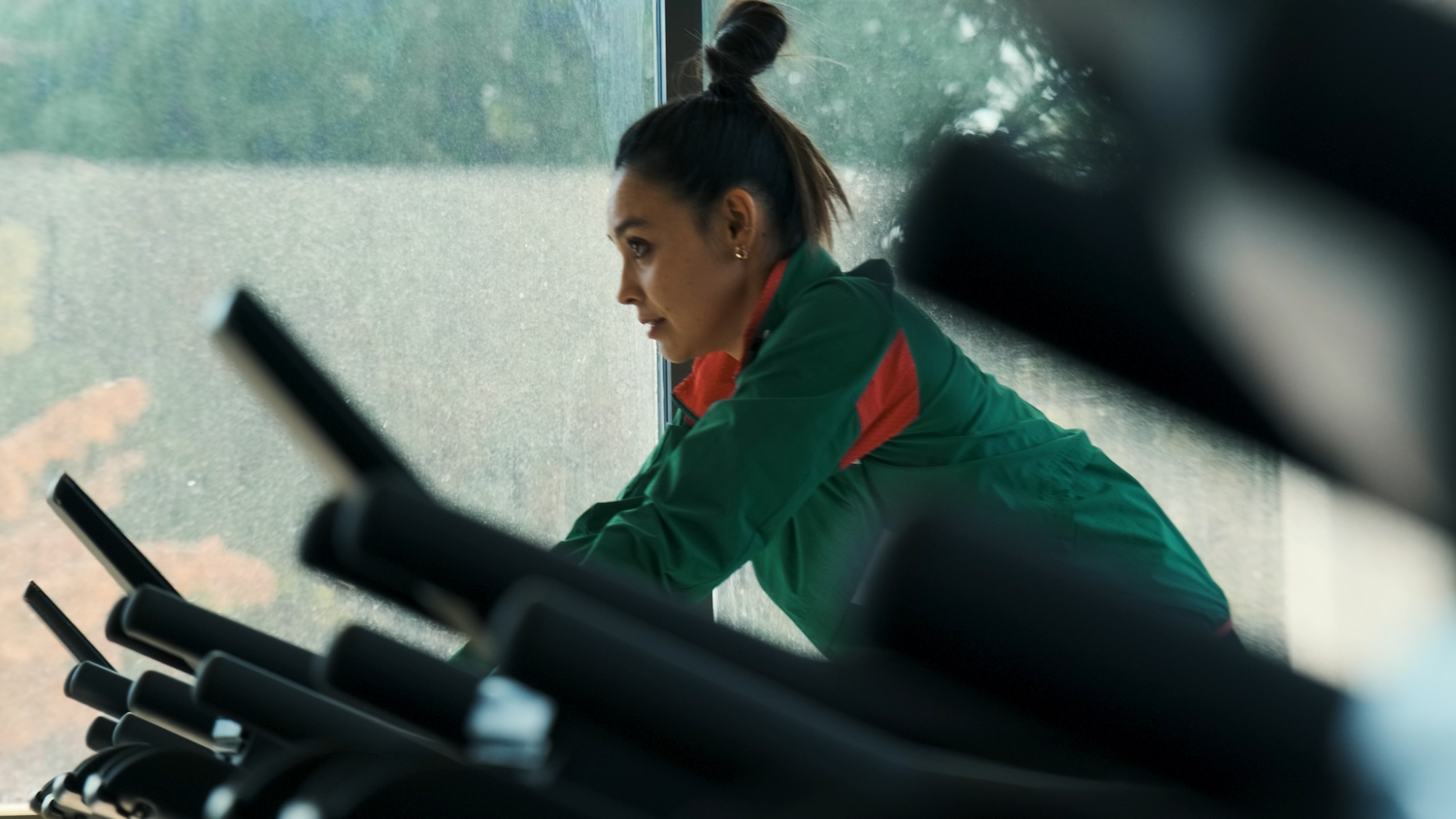 Woman in green jacket exercising on elliptical machine at gym near large window.