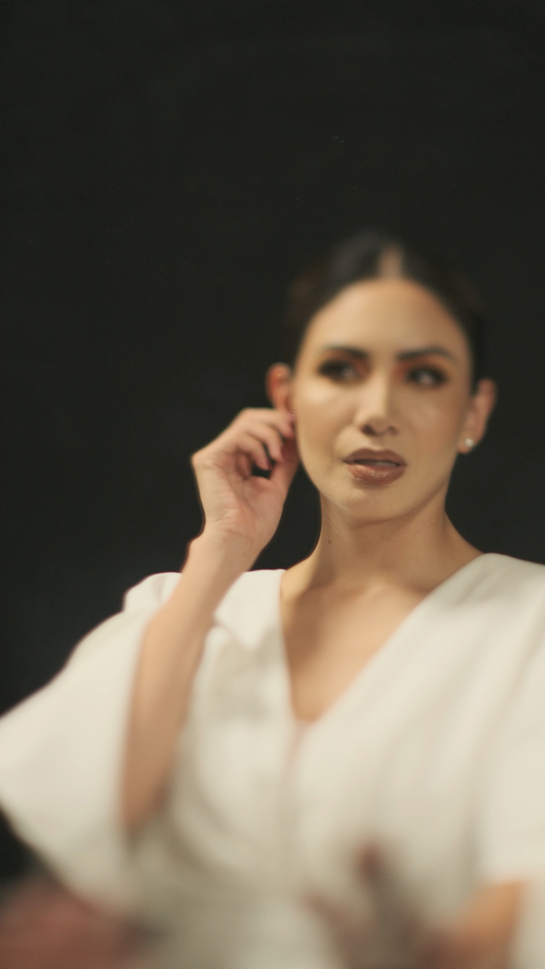 Elegant woman in white dress touches her ear while posing against a dark black background in soft focus.