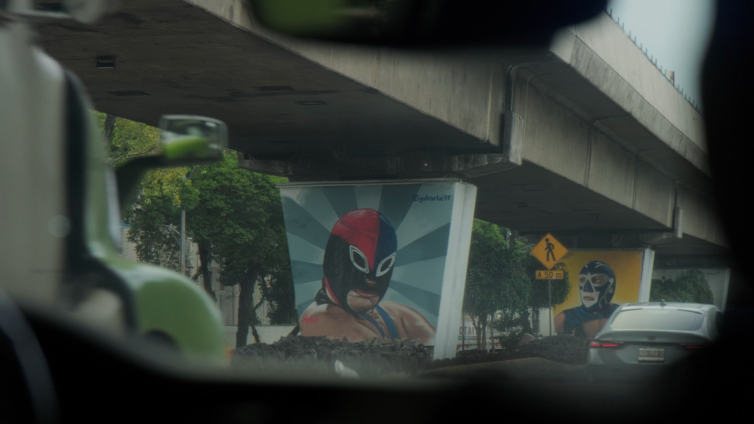 Street mural of a Mexican luchador wrestler mask viewed through a car window in urban traffic.