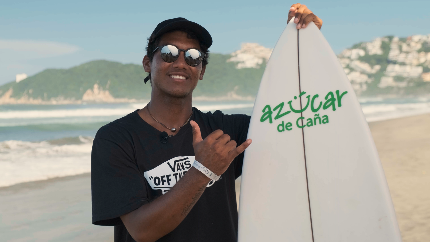 Smiling young man on beach holding a white Azucar de Cana surfboard wearing Vans shirt and cap.