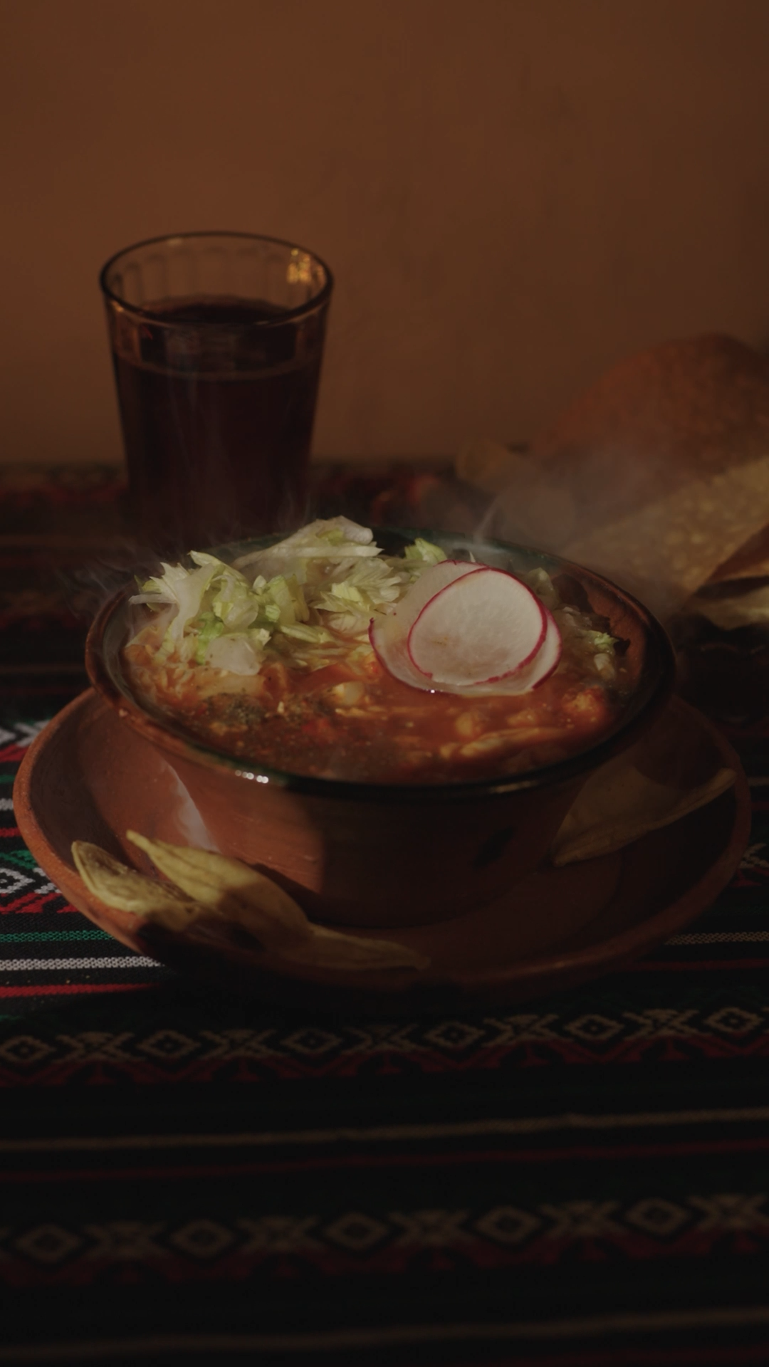 Steaming bowl of pozole with radishes and lettuce on a colorful Mexican tablecloth.