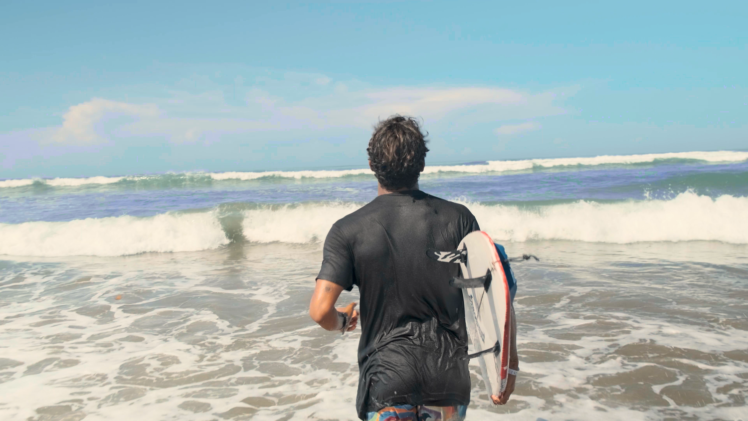 Surfer in black wetsuit walking toward ocean waves carrying a surfboard from behind.