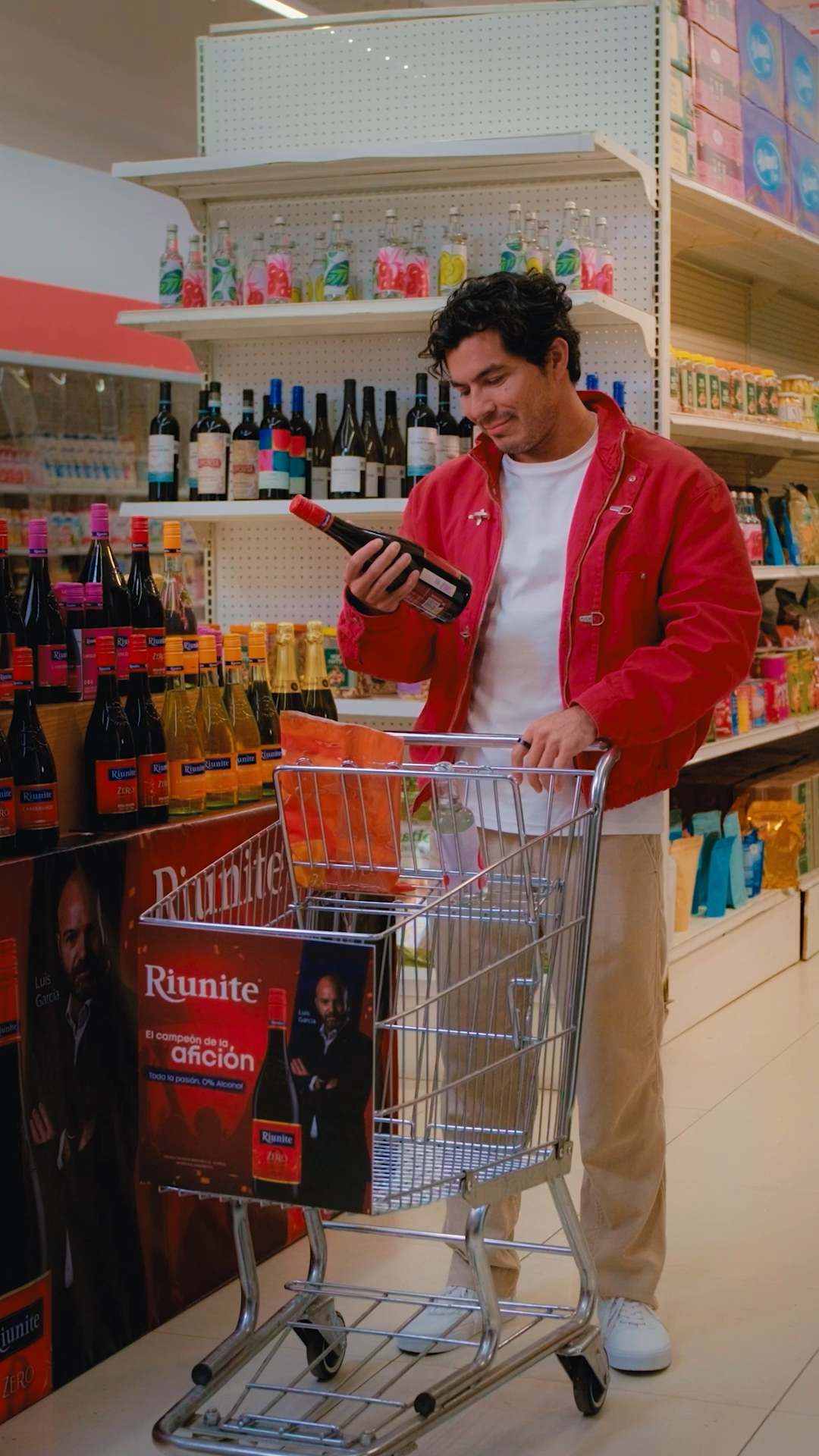 Young man in red jacket examining Riunite wine bottle while pushing shopping cart in grocery store.
