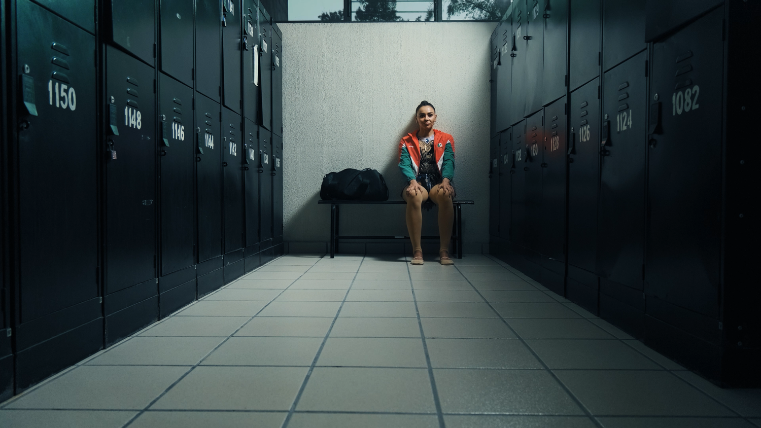 Female athlete in red and blue leotard sitting alone in a locker room on a bench.