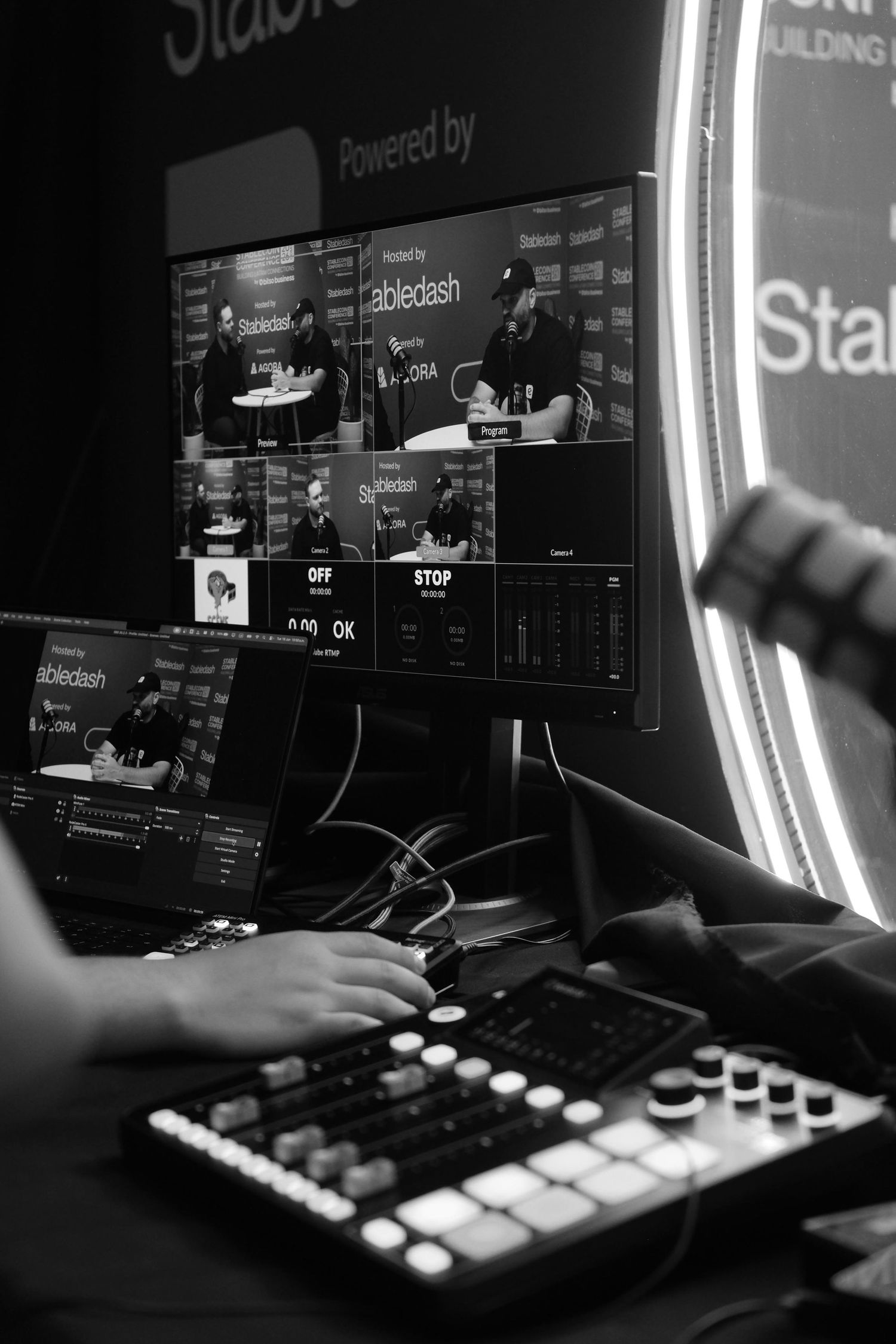 Black and white close-up of hands operating a mixing board with podcast broadcast monitors at Stabledash Conference.