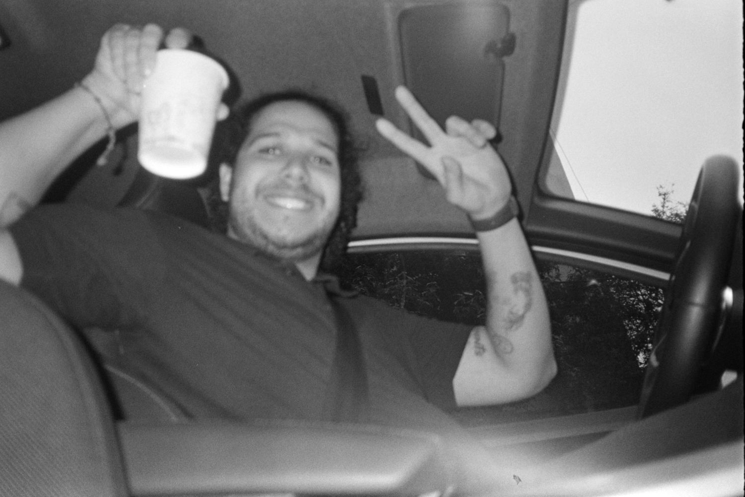 Black and white photo of smiling man in car giving peace sign while holding a drink.
