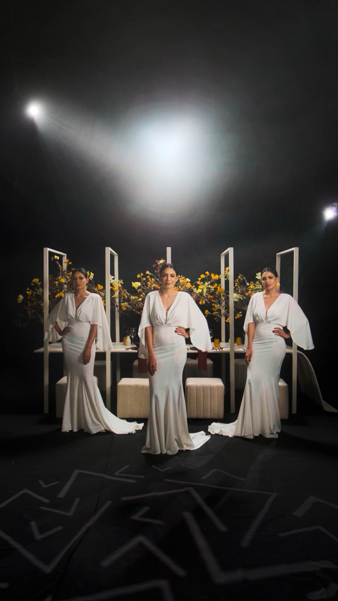 Three women in elegant white gowns pose dramatically under a spotlight with yellow floral arrangements in a dark studio.