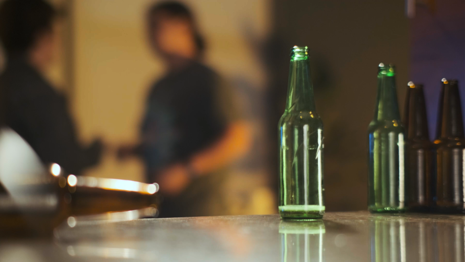 Green glass beer bottles on a bar counter with blurred people conversing in the background.