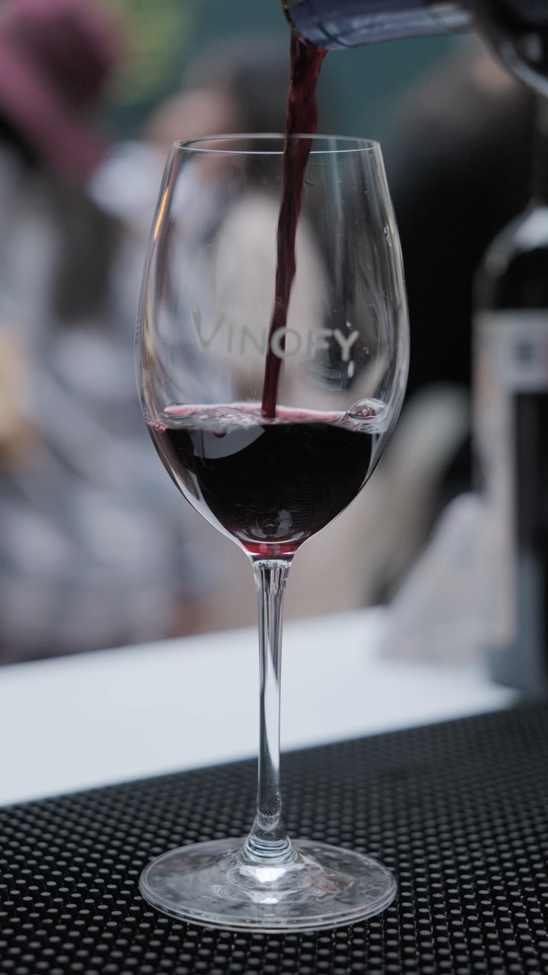 Red wine being poured into Vinofy branded crystal wine glass at tasting event.