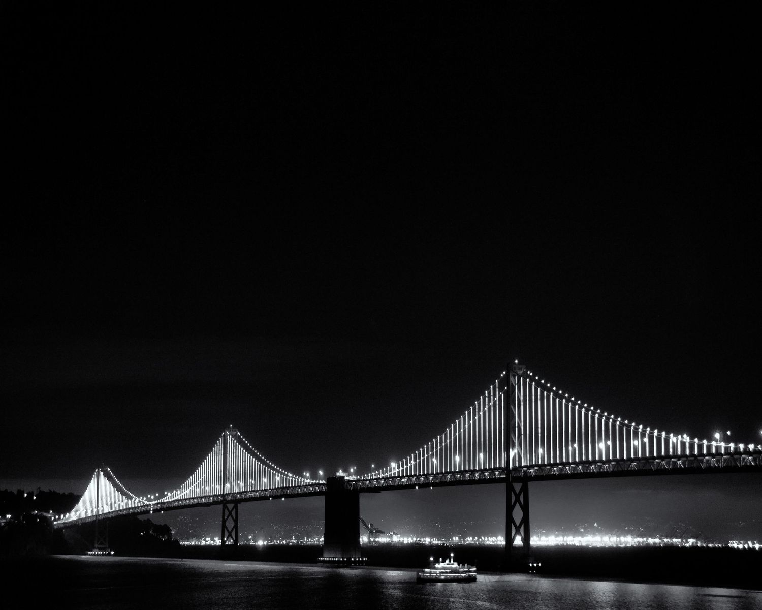 Black and white panoramic nighttime photograph of the San Francisco Bay Bridge with lights reflecting on the bay.