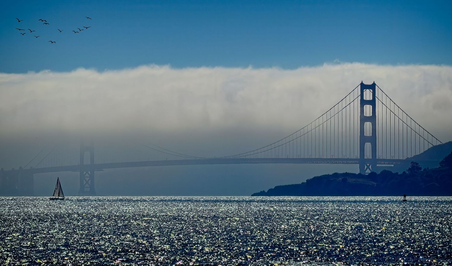 Golden Gate Bridge partially shrouded in morning fog with a sailboat on sparkling San Francisco Bay waters, California, USA.
