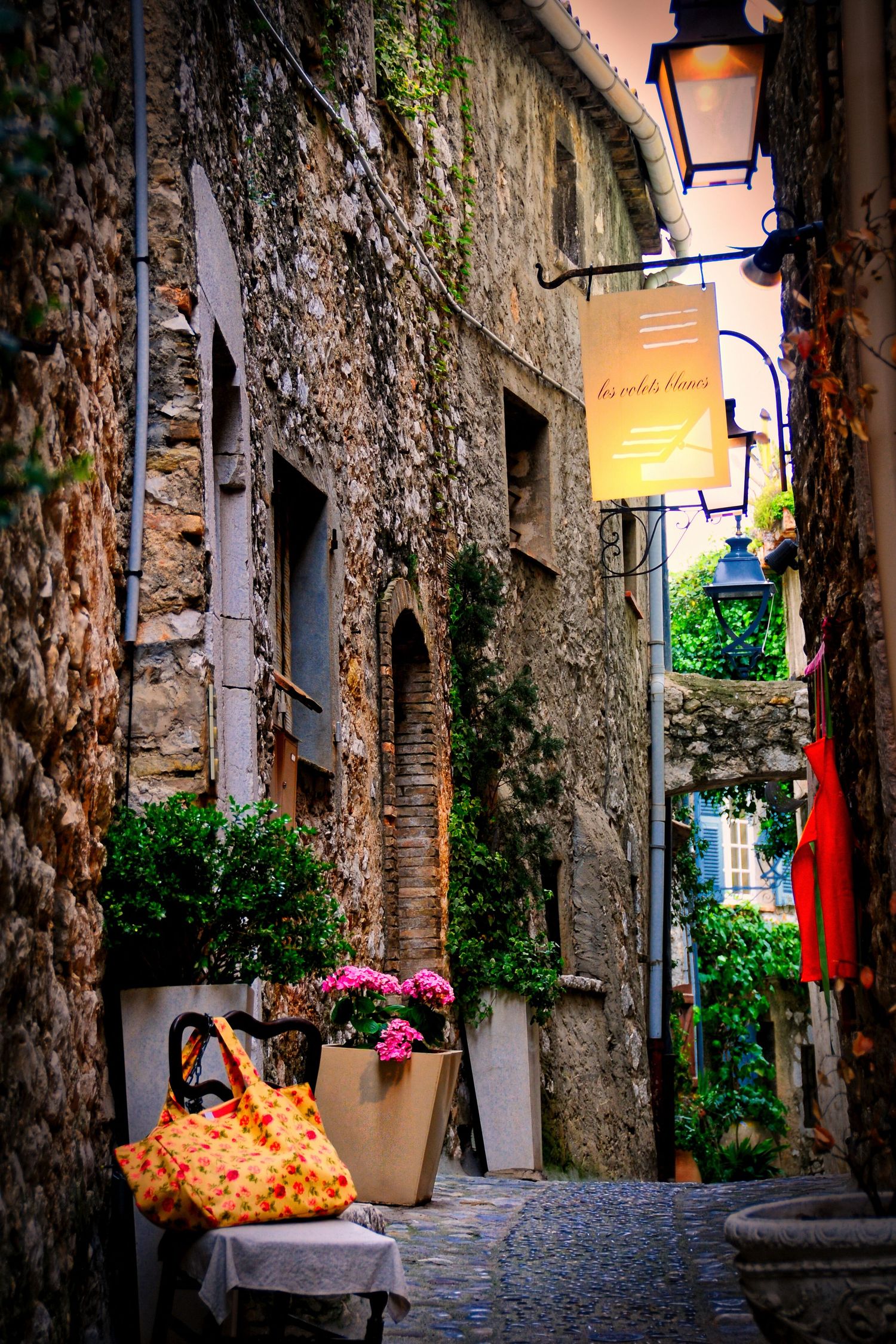 Narrow cobblestone alley in a medieval French village with stone walls and glowing lanterns.
