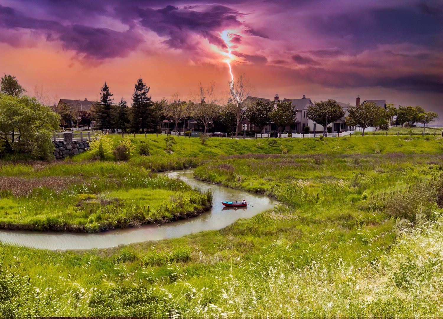 Dramatic lightning strike over a fisherman and lush green wetland landscape with a winding creek and suburban homes at sunset.