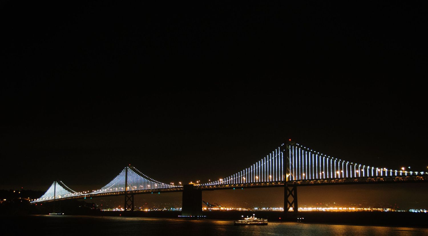 San Francisco Bay Bridge illuminated with brilliant white LED lights reflecting on dark water at night.