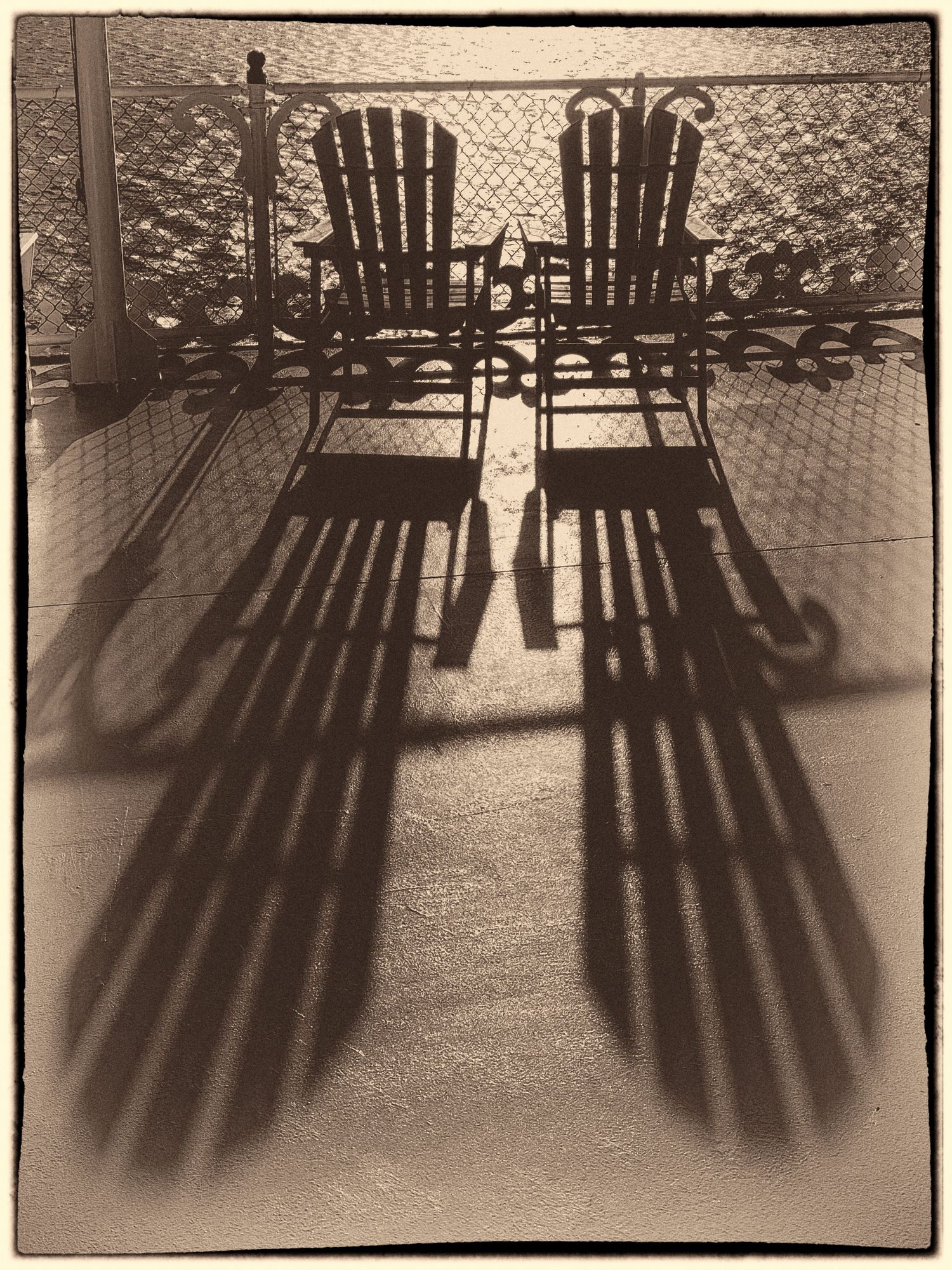 Vintage sepia photo of two Adirondack chairs casting long dramatic shadows on a sunny porch deck.