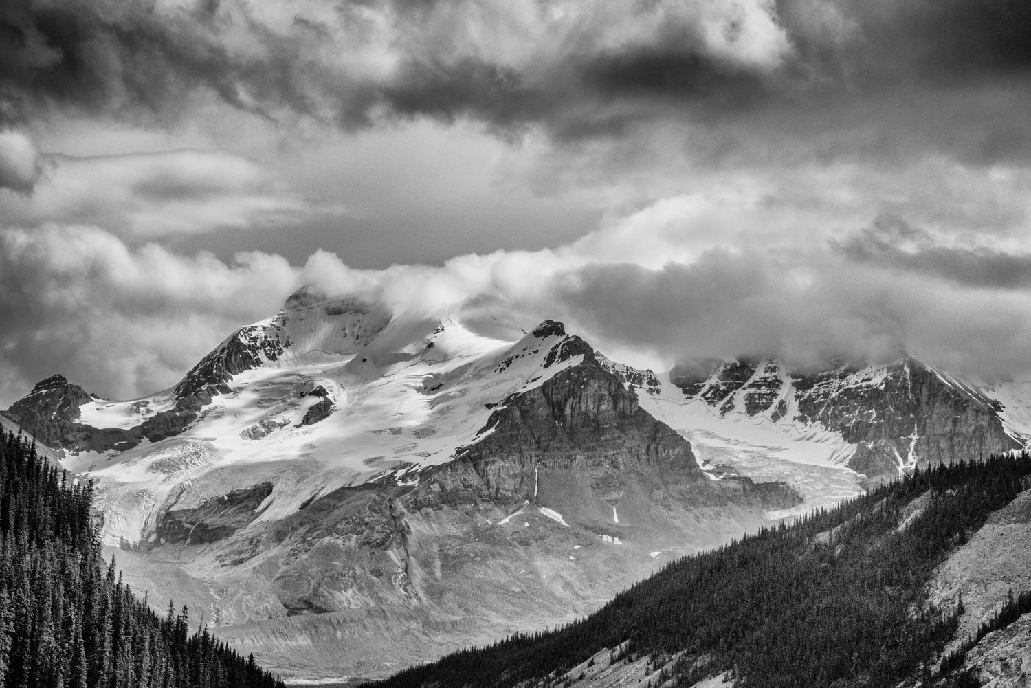 Black and white dramatic mountain landscape with snow-capped peaks and glaciers under stormy clouds.