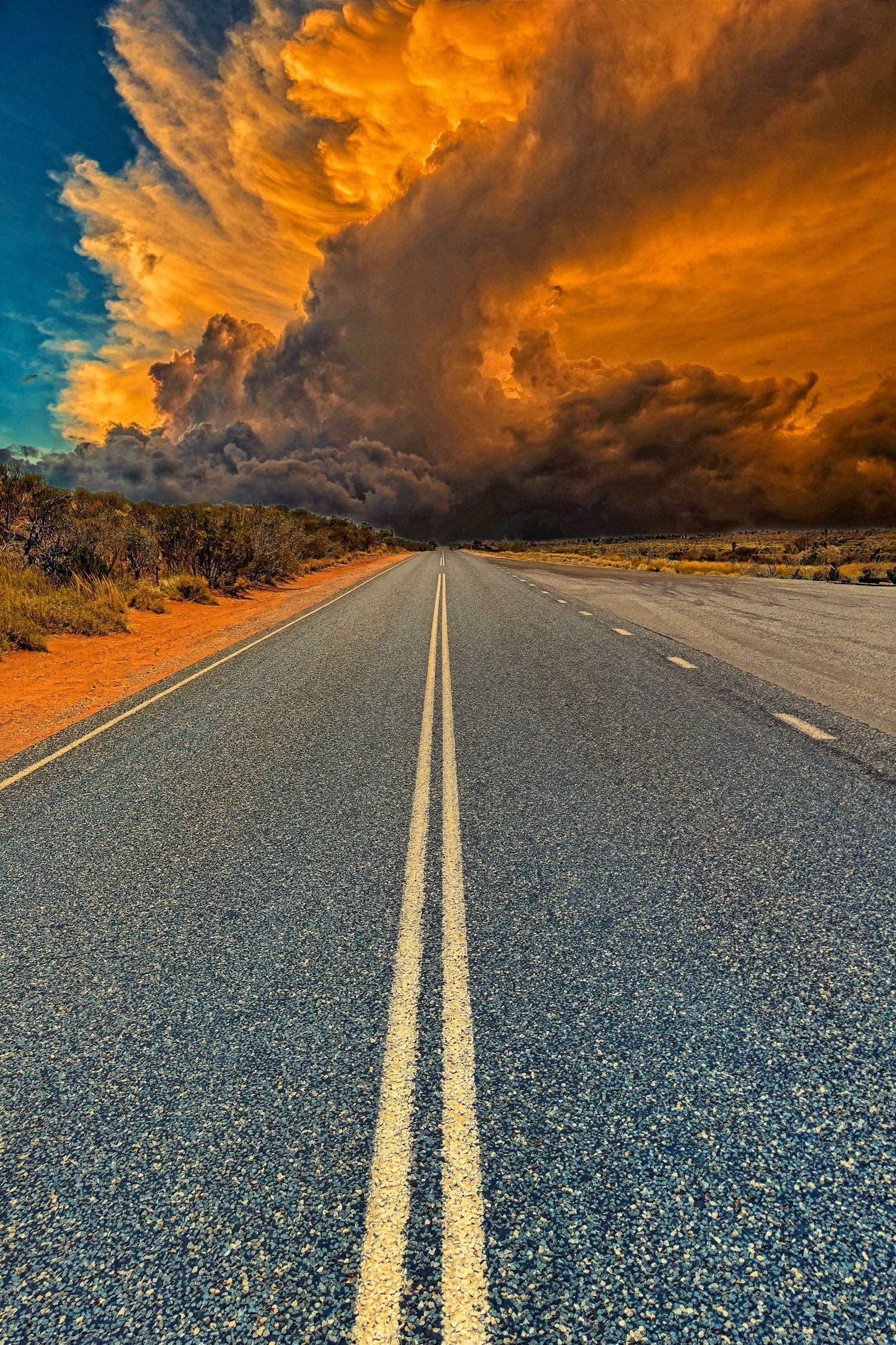 A straight outback road leads toward a massive dramatic storm cloud formation at a vivid orange sunset.