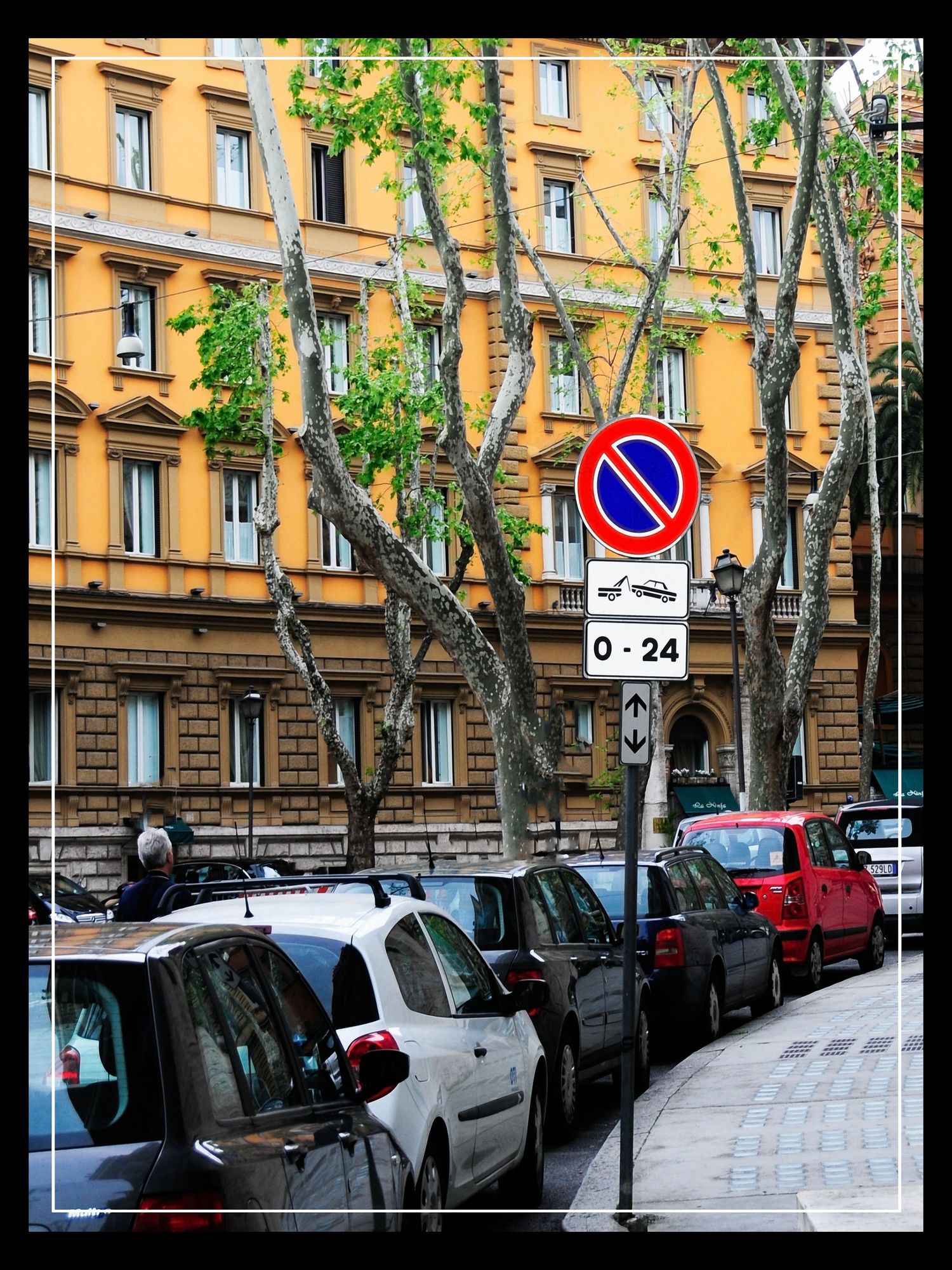 Cars parked along a Roman street with no-parking sign, tall trees, and yellow ochre building facade.