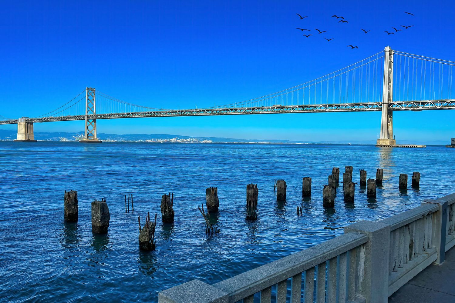 The San Francisco Bay Bridge spans blue water with weathered wooden pilings in the foreground under clear sky.