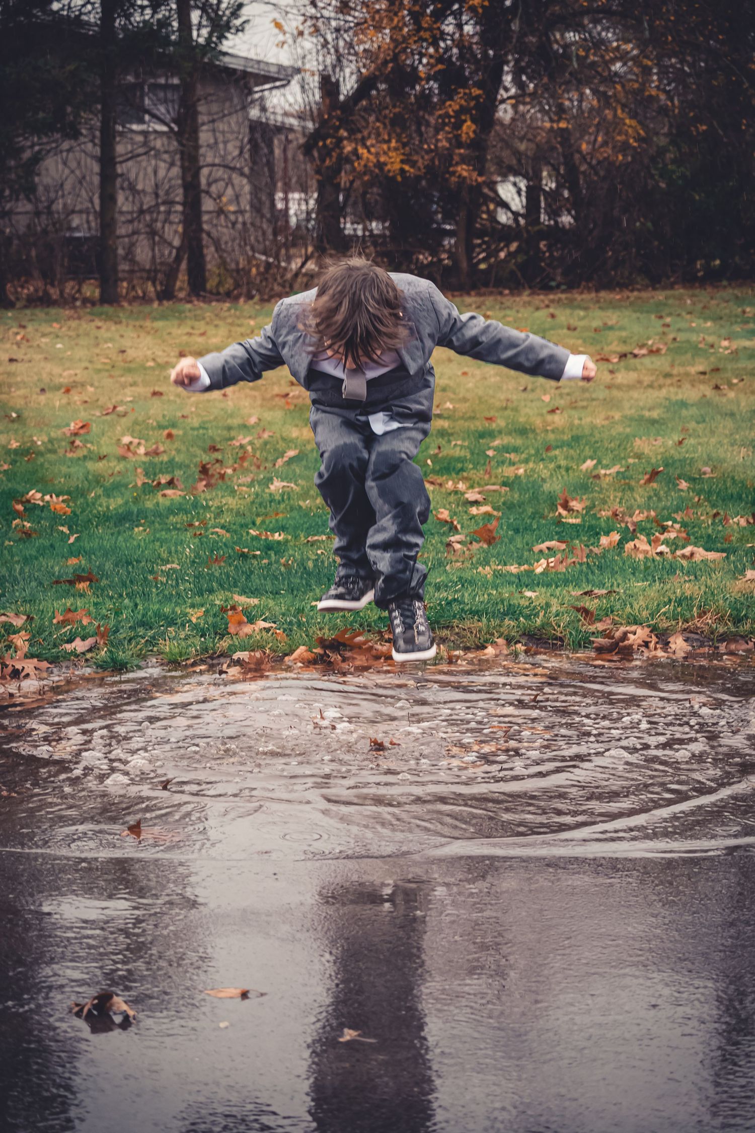 Child jumping into a rain puddle in an autumn park, arms outstretched, surrounded by fallen leaves on green grass.