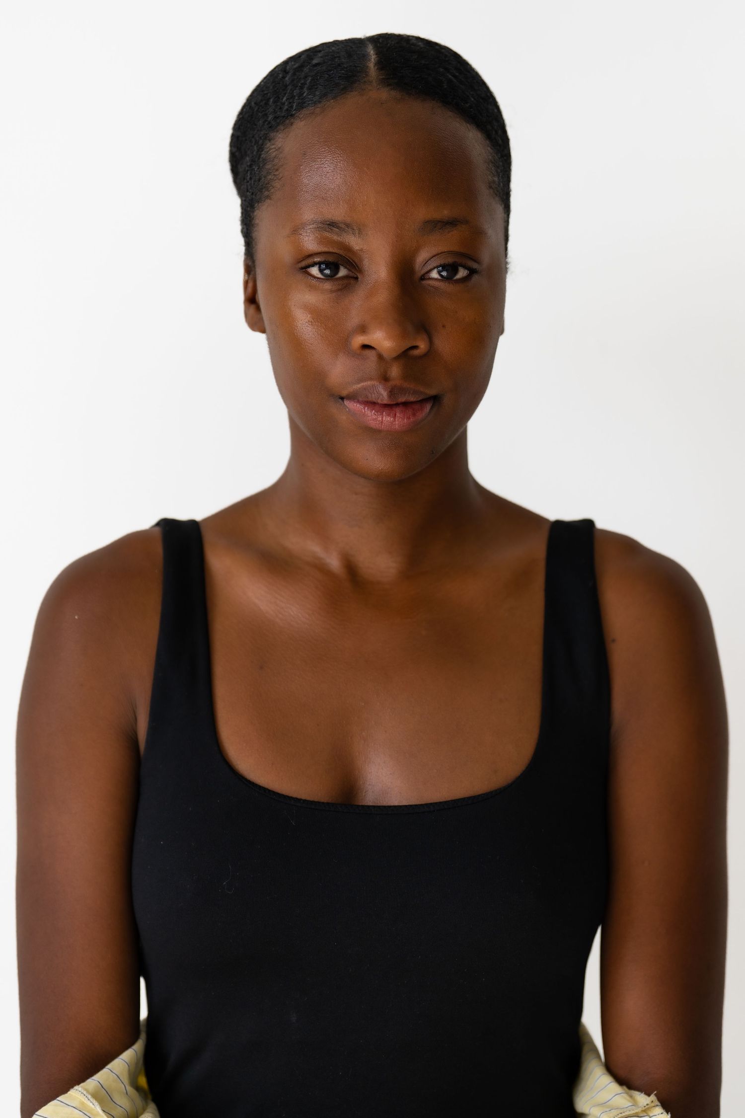 Young Black woman with braided updo in black tank top looking directly at camera against white background.