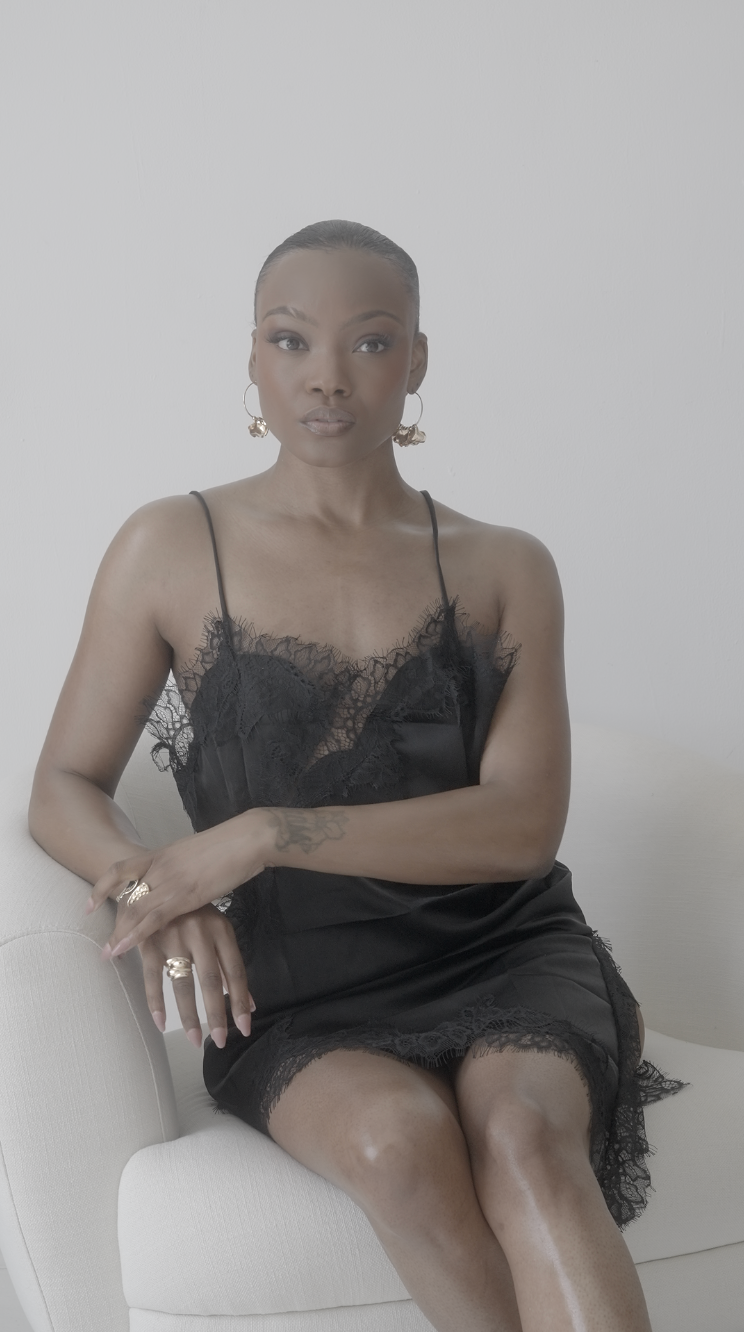 Woman in black lace slip dress seated on white chair, wearing gold hoop earrings and rings, against white background.