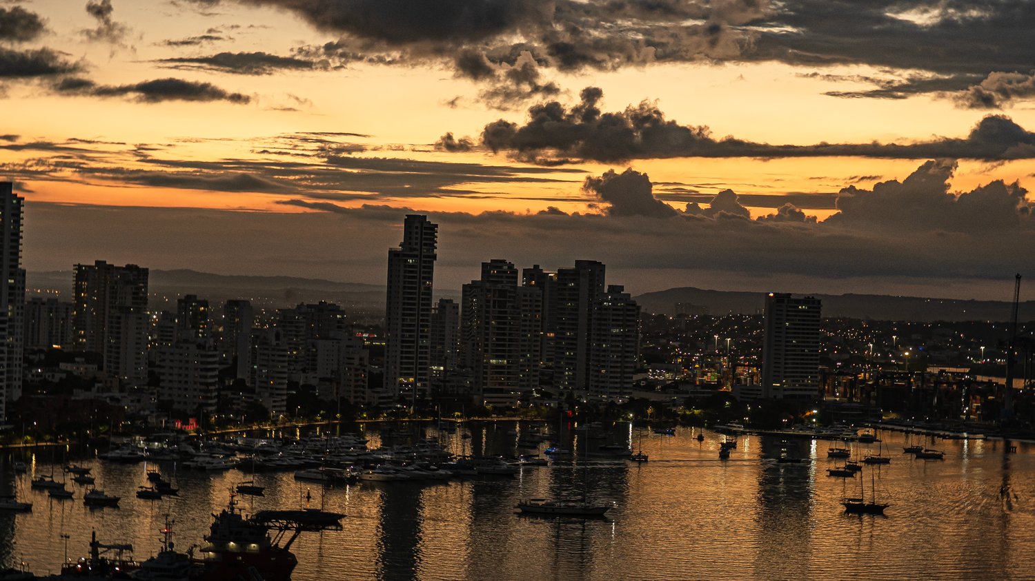 Golden sunset over a city skyline with boats reflected in a harbor under dramatic clouds.