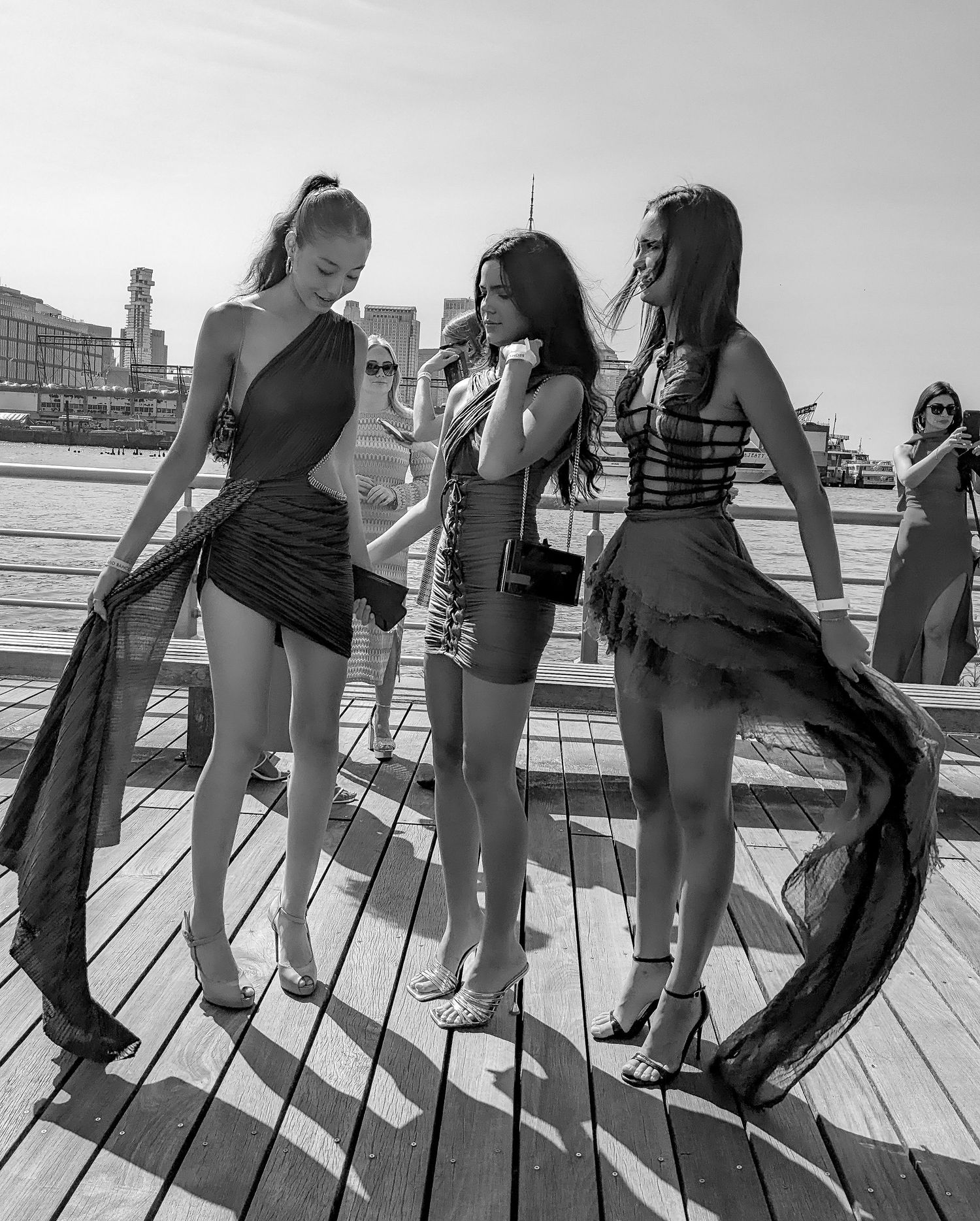 Three stylish women in elegant black dresses pose on a sunny boardwalk in a black and white photo.